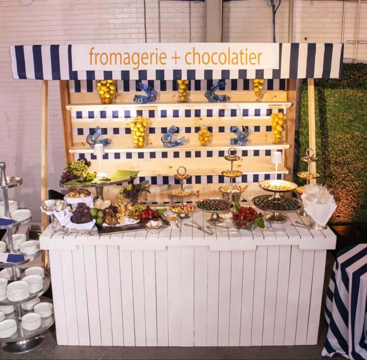 Farmer’s Market Stand measuring 10'L x 4'W x 9'H. Features a white wooden base with slatted design, striped navy-and-white awning, and open shelving for display—perfect for themed food stations, beverage service, or decorative event setups.